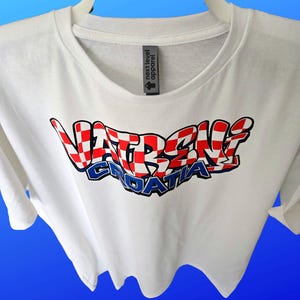 May include: White t-shirt with the text "VATRENI CROATIA" in a red, white, and blue checkered design. The shirt has a crew neck and short sleeves. The brand tag "next level apparel" is visible.