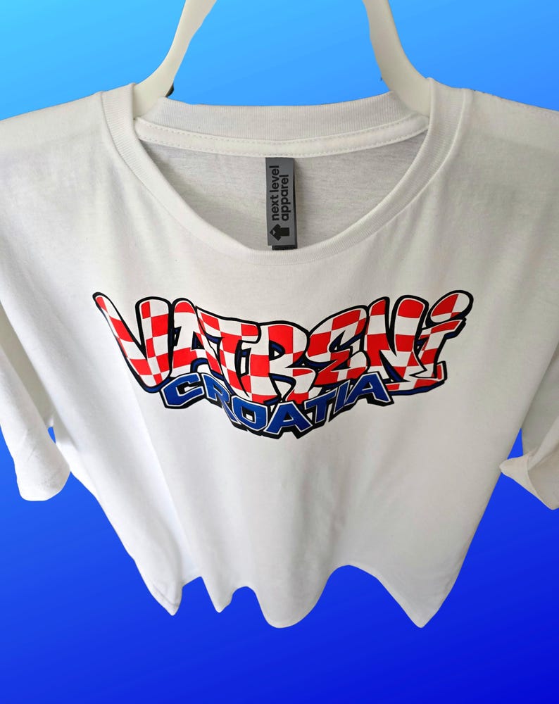 May include: White t-shirt with the text "VATRENI CROATIA" in a red, white, and blue checkered design. The shirt has a crew neck and short sleeves. The brand tag "next level apparel" is visible.