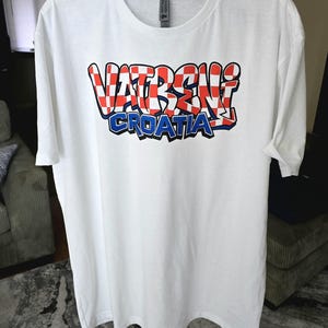 May include: White t-shirt with the text "VATRENI CROATIA" in a red, white, and blue design. The word "VATRENI" is in a checkered pattern, and "CROATIA" is in blue. The shirt is hanging on a black hanger.