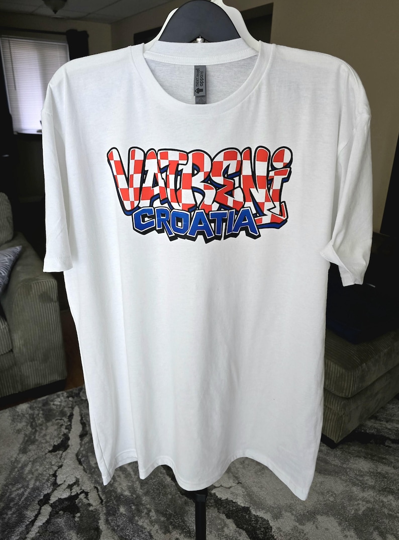 May include: White t-shirt with the text "VATRENI CROATIA" in a red, white, and blue design. The word "VATRENI" is in a checkered pattern, and "CROATIA" is in blue. The shirt is hanging on a black hanger.