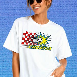 Puede incluir: Una camiseta blanca de manga corta con un estampado gr&aacute;fico de un bal&oacute;n de f&uacute;tbol atravesando un muro a cuadros rojos y blancos. El dise&ntilde;o incluye un efecto de estrella amarillo y azul y la palabra CROATIA en letras amarillas.