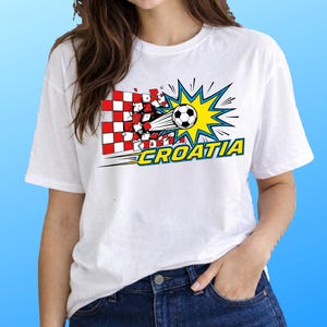 Puede incluir: Una camiseta blanca de manga corta con un gr&aacute;fico de un bal&oacute;n de f&uacute;tbol atravesando una pared de cuadros rojos y blancos. El dise&ntilde;o incluye un efecto de estallido amarillo y la palabra CROATIA en letras amarillas con borde azul.