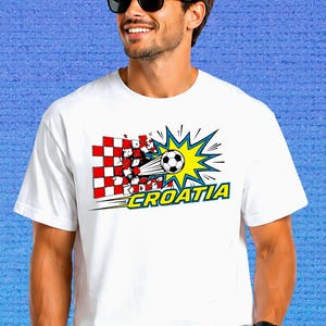 Puede incluir: Una camiseta blanca de manga corta con un estampado gr&aacute;fico de un bal&oacute;n de f&uacute;tbol atravesando una pared de cuadros rojos y blancos. El dise&ntilde;o incluye un efecto de estallido amarillo y la palabra CROATIA en letras amarillas con bordes azules.