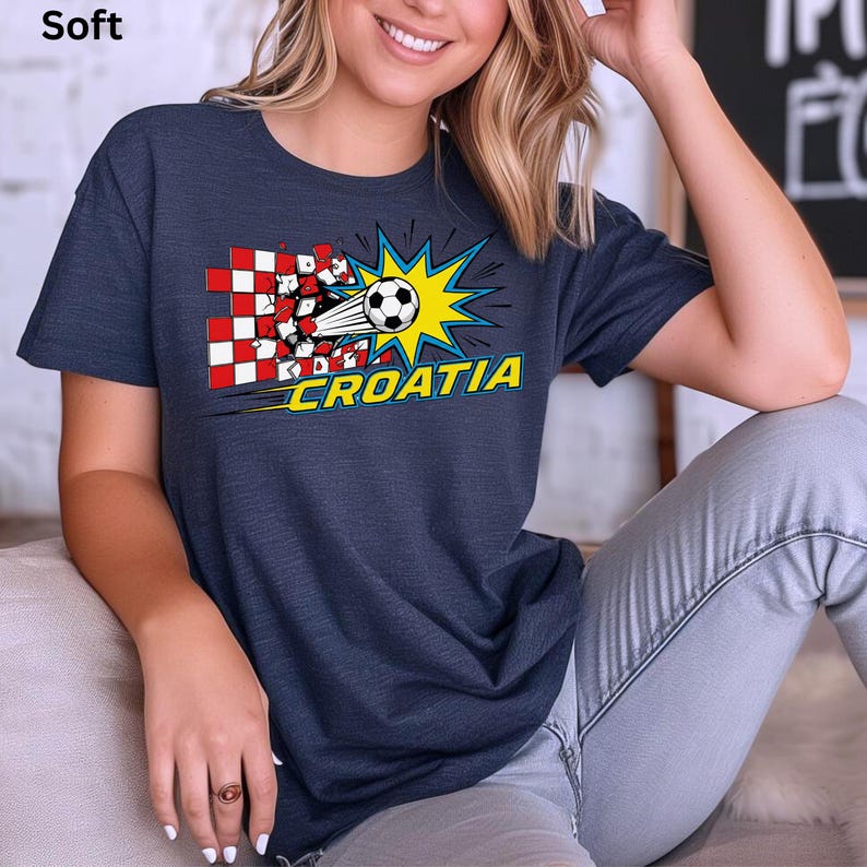 Op de afbeelding: Marineblauw t-shirt met een voetbal die door een rood-wit geblokte vlag breekt. Het woord "CROATIA" is geel met een blauwe omlijning. Het shirt heeft een zachte textuur.