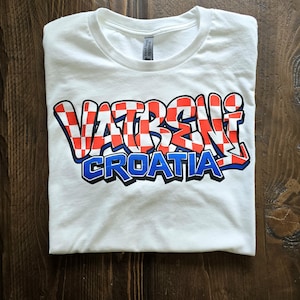 May include: White t-shirt with the words "VATRENI CROATIA" in a red and white checkered design with blue and black accents. The shirt is folded on a wooden surface.