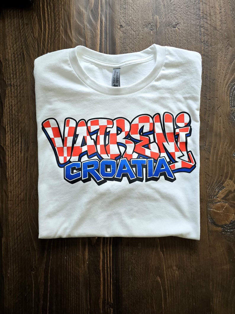 May include: White t-shirt with the words "VATRENI CROATIA" in a red and white checkered design with blue and black accents. The shirt is folded on a wooden surface.