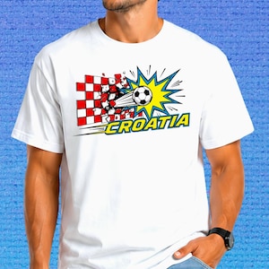 Puede incluir: Una camiseta blanca de manga corta con un estampado gr&aacute;fico de un bal&oacute;n de f&uacute;tbol atravesando una pared de cuadros rojos y blancos. El dise&ntilde;o incluye un efecto de estallido amarillo y la palabra CROATIA en letras amarillas con borde azul.