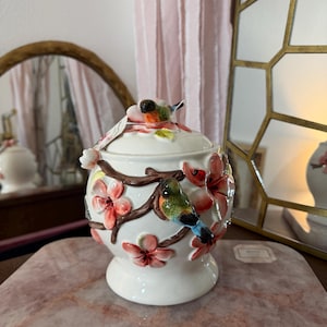 May include: A white ceramic jar with a lid, adorned with colorful birds and pink floral accents. The jar features a round shape with a pedestal base. The birds are perched on brown branches, and the flowers are in full bloom.