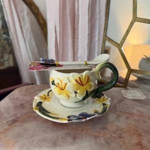 May include: A decorative teacup and saucer set with a matching spoon. The cup and saucer are cream-colored with yellow and white floral designs and green accents. The spoon is white with a floral design on the handle.