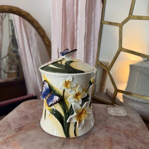 May include: A decorative ceramic jar with a lid, featuring a butterfly and floral design. The jar is cream-coloured with hand-painted details of blue butterflies and white daffodils. The lid is adorned with a butterfly. The jar is approximately 15 cm tall.