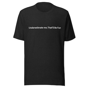 Op de afbeelding: Donkergrijze t-shirt met de witte tekst "Underestimate me. That'll Be Fun". Het shirt heeft een ronde hals en korte mouwen. De stof heeft een gemêleerde textuur.