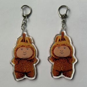 May include: Two keychains featuring a cartoon character in a brown furry costume with a yellow hood. The character has a round face and a neutral expression. Each keychain has a silver-colored metal ring.