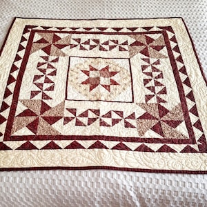 May include: A handmade quilt with a central star design in shades of burgundy and cream. The quilt features geometric patterns, including triangles and pinwheels, and is bordered by a burgundy trim. The quilt is approximately 152 cm by 152 cm.