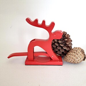 May include: A bright red metal nutcracker shaped like a stylised reindeer with a lever handle and a textured gripping base. The nutcracker is positioned next to two brown pinecones, one large and one smaller, on a plain white surface.