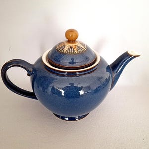 May include: A blue ceramic teapot with a matching lid. The teapot has a rounded body, a curved handle, and a spout. The lid is topped with a small, round, wooden knob. The teapot is glazed with a glossy finish.
