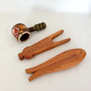 May include: A collection of three wooden kitchen tools including a dark brown pipe-shaped nutcracker with hand-painted floral details, a light brown nutcracker with intricate geometric carvings, and a fish-shaped wooden nutcracker tool.