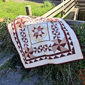 May include: A square quilt featuring a central star block surrounded by a border of pinwheel and triangle patterns. The design uses a colour palette of deep maroon and cream, with intricate stitching visible across the entire fabric surface.