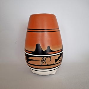 May include: A hand-painted ceramic vase with a Southwestern design. The vase features a terracotta-coloured body with black and white geometric patterns and a landscape scene. The vase is approximately 18 cm tall.