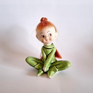 May include: A vintage ceramic figurine of a seated figure with a green outfit and red hair. The figurine has a smiling face and is in a cross-legged position. The figure is wearing a green outfit with a high collar.