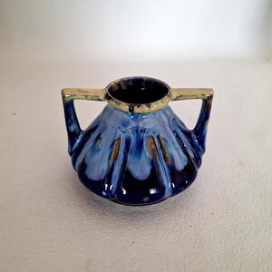 May include: A small ceramic vessel featuring two handles and a wide, fluted base. The exterior is finished with a glossy glaze in shades of deep navy blue, lighter sky blue, and accents of warm tan, particularly around the rim and handle tops.
