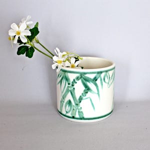 May include: A white ceramic vase with a green bamboo design. The cylindrical vase holds a sprig of white flowers with yellow centres and green leaves. The vase has a green border at the top and bottom.