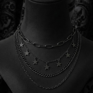 Peut inclure: Ensemble de colliers noirs superposés présenté sur un buste de mannequin noir. Les colliers présentent différents styles de chaînes, dont une chaîne épaisse et des chaînes avec des pendentifs en forme d'étoiles. Le fond est un tissu sombre à motifs.