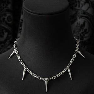 May include: A silver-toned necklace with a chain design and multiple cone-shaped spikes. The necklace is displayed on a black mannequin bust against a dark patterned background. The jewelry is a gothic or punk style.
