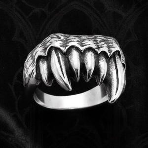 May include: A silver-toned metal ring featuring a sculpted design of a row of sharp, pointed teeth or fangs. The band is smooth and circular, while the top portion displays textured, jagged details resembling a creature's mouth.