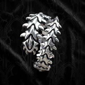 May include: A silver-toned metal ring featuring a segmented, articulated design that resembles a spinal column or vertebrae. The surface has a polished finish with deep, dark recessed areas that highlight the intricate, repeating bone-like shapes.