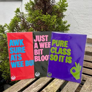 Può includere: Tre stampe artistiche colorate con testo in grassetto. La stampa rossa dice "AWK SURE ATS WEE BU" in blu. La stampa nera dice "JUST A WEE BIT BLOO'S" in rosa. La stampa viola dice "PURE CLASS SO IT IS" con un gesto della mano verde.