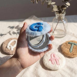 May include: A compact mirror with a light blue fluffy cover and a blue embroidered "G". Several round, fluffy coasters with embroidered letters are also visible. The items are on a cream-colored surface.