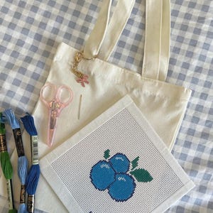 May include: A cream-colored tote bag with a pink bow charm, pink scissors, and embroidery supplies. A cross-stitch project features a blueberry design with the text "NP CLUB". Blue and green embroidery floss is also included.