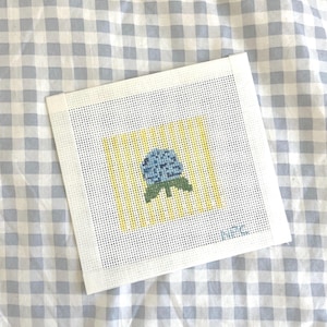 May include: Square needlepoint canvas featuring a blue flower with green leaves, set against yellow vertical stripes. The canvas is on a white background with a gingham pattern in the background.