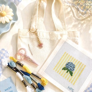 May include: A cream-colored tote bag with a gold keychain and pink charm, surrounded by embroidery supplies. Includes pink scissors, embroidery floss in blue, yellow, and white, and a needlepoint canvas with a floral design. A small card reads "MADE WITH".