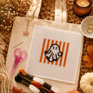 May include: A beige canvas tote bag featuring a square cross-stitch project with a white ghost on orange vertical stripes. The set includes a pink pair of scissors, a needle, and three skeins of embroidery floss in black, orange, and white colors.