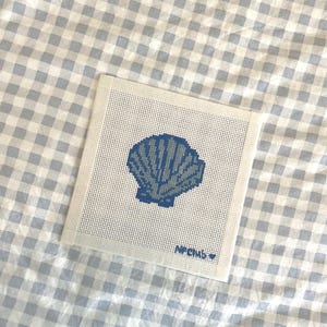 May include: Square needlepoint canvas featuring a blue seashell design. The seashell is stitched in a pixelated style, with a darker blue outline and lighter blue fill. The text "NPCMB" is stitched in the bottom right corner.