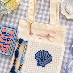 May include: A cream-colored tote bag with a gold charm and pink bow, next to a cross-stitch project of a blue seashell. Also includes blue embroidery floss, pink scissors, and a sardine tin with a fish graphic.