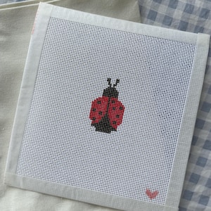May include: A square needlepoint canvas featuring a red and black ladybug design. The ladybug is centered on a white mesh background, with a small pink heart in the corner. The canvas is bordered by a white fabric and rests on a light beige cloth.