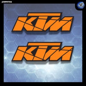 Peut inclure: Deux logos KTM orange avec des contours noirs et des lignes géométriques noires internes. Le texte STORMSTICK apparaît dans le coin supérieur gauche, et un logo circulaire avec le texte STORMSTICK est situé dans le coin supérieur droit.
