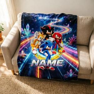 Puede incluir: Una manta de forro polar rectangular con un fondo de temática espacial, cristales brillantes y estelas de luz coloridas. El diseño muestra a los personajes Sonic, Tails, Shadow y Knuckles sobre la palabra NAME en letras blancas.