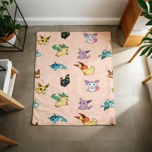 May include: A rectangular fleece blanket in a soft peach color featuring a repeating pattern of various colorful cartoon creatures, including yellow, blue, purple, and black characters with distinct ears and tails, scattered across the fabric.