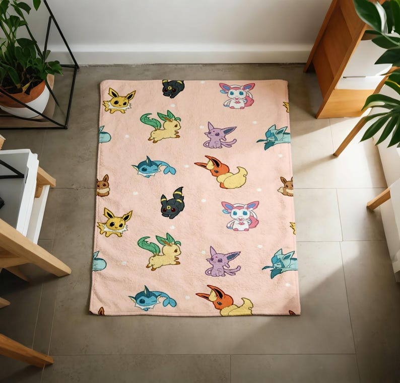 May include: A rectangular fleece blanket in a soft peach color featuring a repeating pattern of various colorful cartoon creatures, including yellow, blue, purple, and black characters with distinct ears and tails, scattered across the fabric.