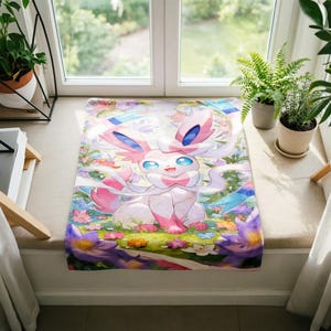 May include: A rectangular fabric mat featuring a colorful illustration of the character Sylveon. The creature is light pink and white with large blue eyes, surrounded by a vibrant garden of purple, yellow, and white flowers on a green background.