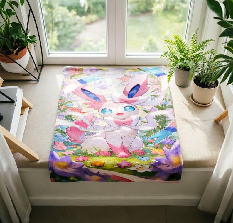 May include: A rectangular fabric mat featuring a colorful illustration of the character Sylveon. The creature is light pink and white with large blue eyes, surrounded by a vibrant garden of purple, yellow, and white flowers on a green background.