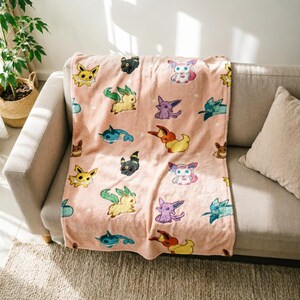 May include: A soft, light pink fleece throw blanket featuring a repeating pattern of various colorful cartoon creatures, including yellow, blue, purple, and brown characters with distinct ears and tails, arranged across the fabric surface.