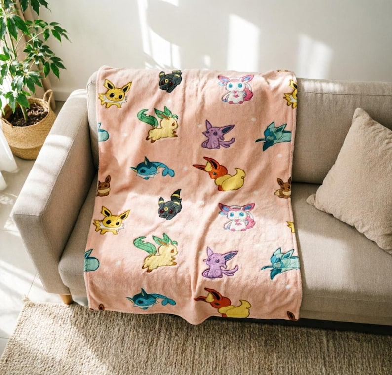 May include: A soft, light pink fleece throw blanket featuring a repeating pattern of various colorful cartoon creatures, including yellow, blue, purple, and brown characters with distinct ears and tails, arranged across the fabric surface.