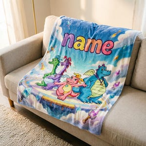 Puede incluir: Una manta azul suave con una ilustración de tres dragones. La manta tiene un fondo de cielo y la palabra "name" en colores arcoíris. Los dragones son en tonos de verde, morado y rosa, con un tema de fantasía.