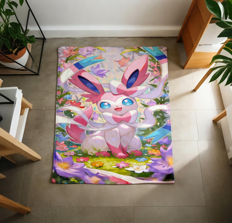 May include: A rectangular area rug featuring a colorful illustration of the pink and white Sylveon character from the Pok&eacute;mon franchise, surrounded by a vibrant garden of purple, orange, and white flowers and green foliage on a light background.