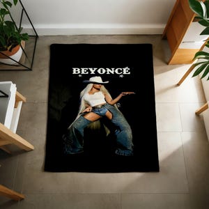 May include: Black rectangular throw blanket featuring a photograph of Beyoncé in a white top, denim chaps, and a white cowboy hat. The word "BEYONCÉ" is printed in white above the image, with stars on either side.