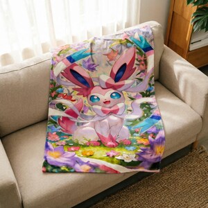 May include: A soft throw blanket featuring a colorful illustration of the pink and white Sylveon character from the Pok&eacute;mon franchise, surrounded by a vibrant garden of purple, yellow, and white flowers with green foliage and blue ribbons.